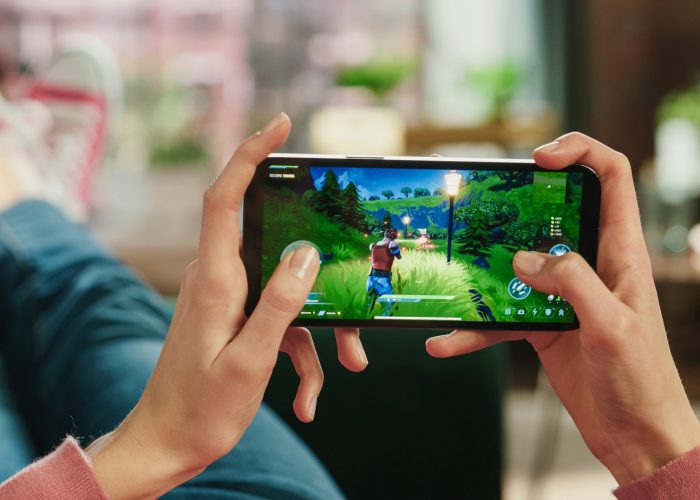 Female is Relaxing on a Couch at Home, Playing an Interactive PvP Shooter Video Game on Her Smartphone. Gamer Lies on a Sofa in Living Room. Close Up POV Photo of Mobile Device Screen.